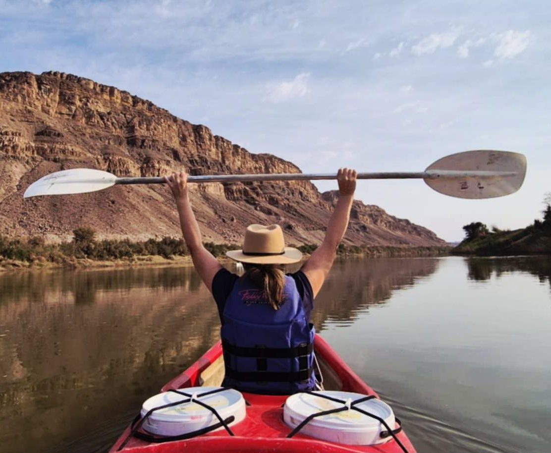 Canoer or rafter with paddle raised in victory or happy stance above head as she floats down the Orange River with mountain to the side