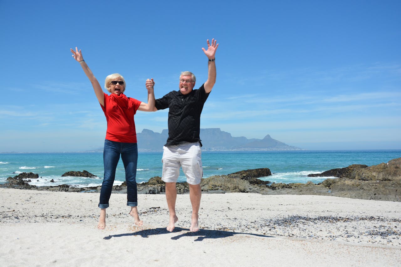 Senior couple holding hands and jumping in air on beach with Table Mountain in distance across the bay