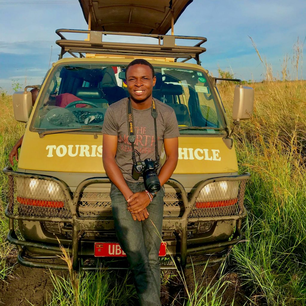 Smiling Ugandan guide with binos around neck leaning back against his safari behicle