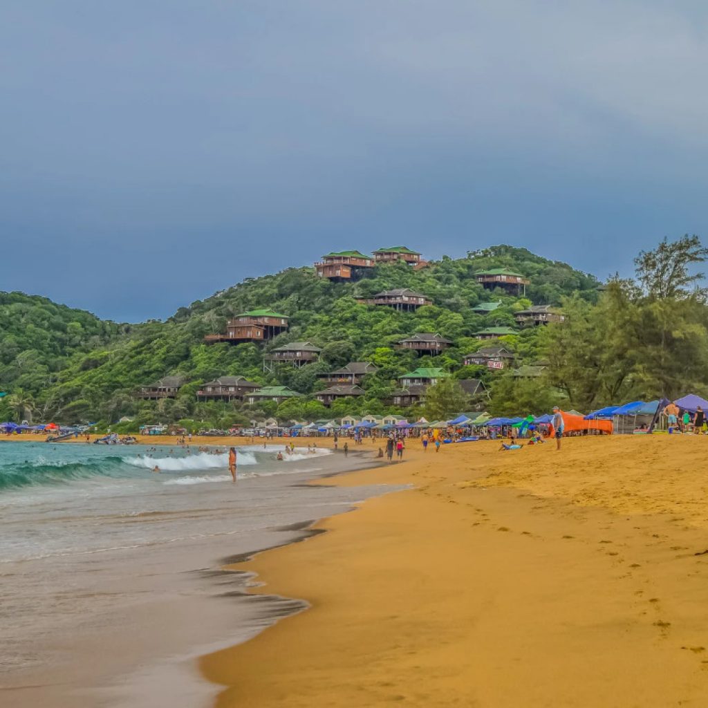 Sandy beach of Ponta Do Ouro with cabins on shrub-covered dunes and people on the beach, Explore Mozambique