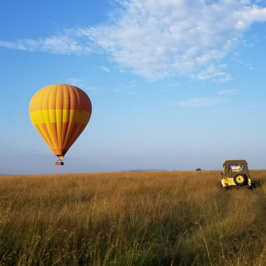 A yellow and orange hot air balloon ascending over the golden savanna of the Maasai Mara, Kenya, with an open safari vehicle parked on the plains below.