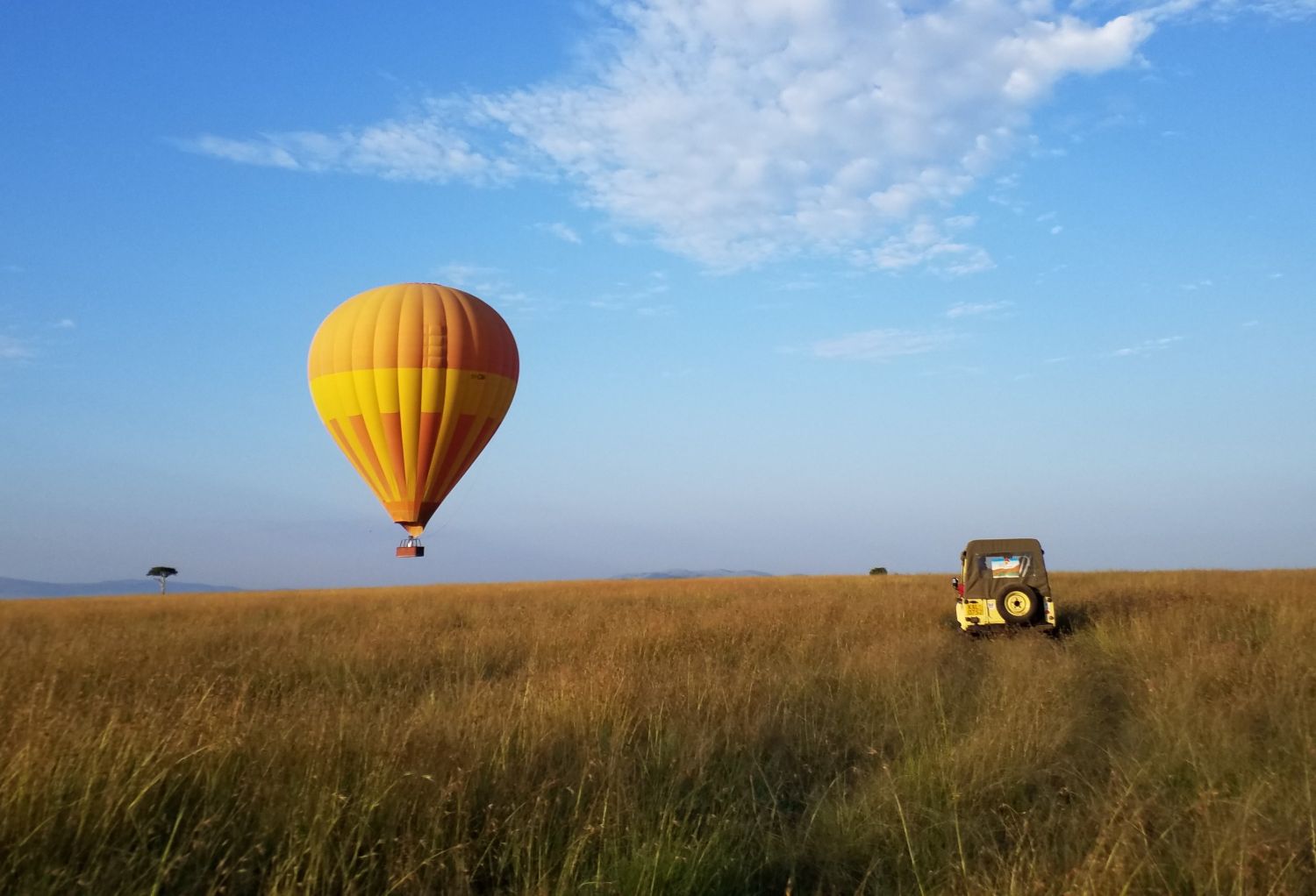 A yellow and orange hot air balloon ascending over the golden savanna of the Maasai Mara, Kenya, with an open safari vehicle parked on the plains below.