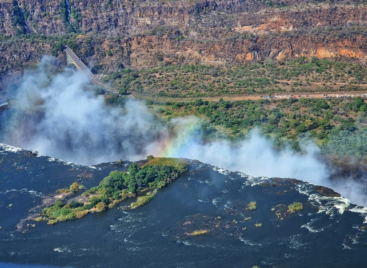Aerial view of the mighty Victoria Falls from above, showing the Zambezi River cascading over the edge, creating mist and a rainbow, with the Victoria Falls Bridge and lush landscape of Zambia and Zimbabwe in the background.