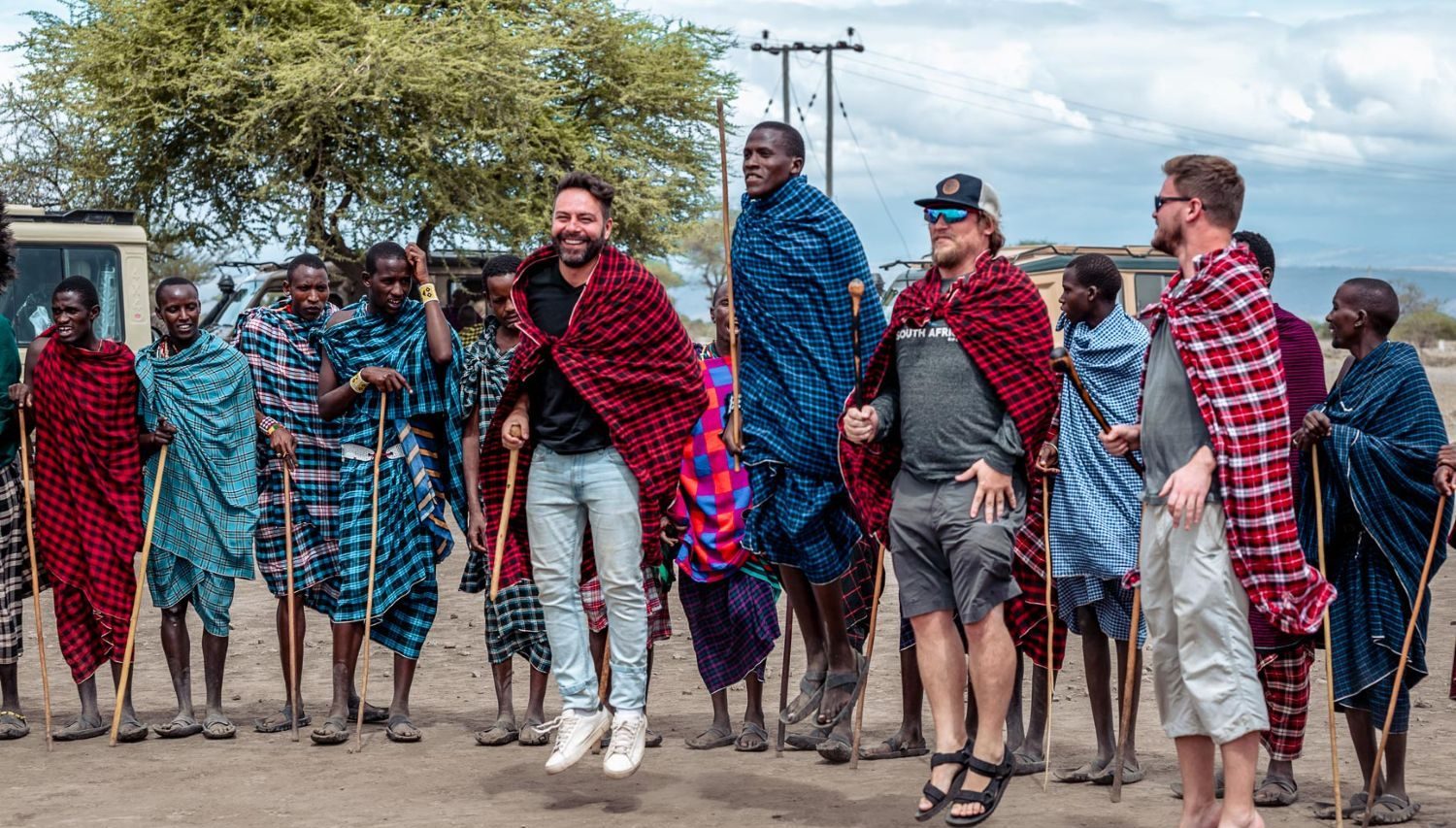 SEASONED TRAVELERS: Tired of tourist traps? Our overland tours offer genuine, immersive cultural exchanges, allowing you to ditch the guidebook and participate in local traditions, like learning the iconic adumu jump with the Maasai.Two seasoned male travelers jumping alongside a group of Maasai warriors in traditional red and blue shukas during a cultural visit in Tanzania or Kenya.
