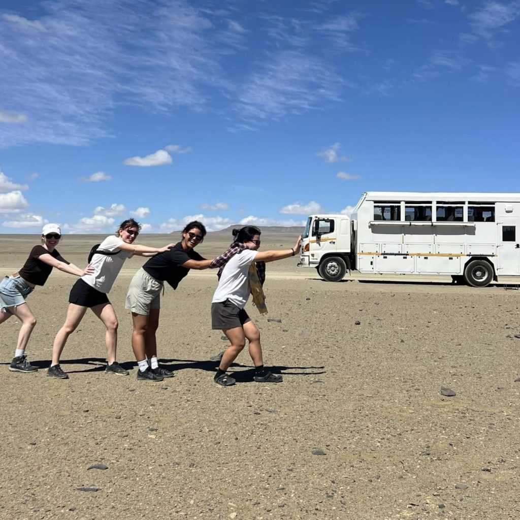 Four friends playfully posing against their white overland truck on a vast, arid plain in Namibia, enjoying a shared adventure and creating fun travel memories.