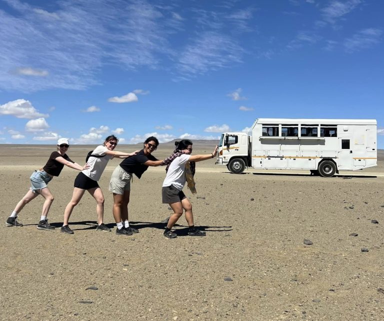 Four friends playfully posing against their white overland truck on a vast, arid plain in Namibia, enjoying a shared adventure and creating fun travel memories.
