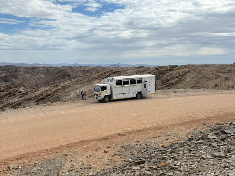 A wide panoramic view of an overland safari truck driving through a vast and beautiful African landscape.