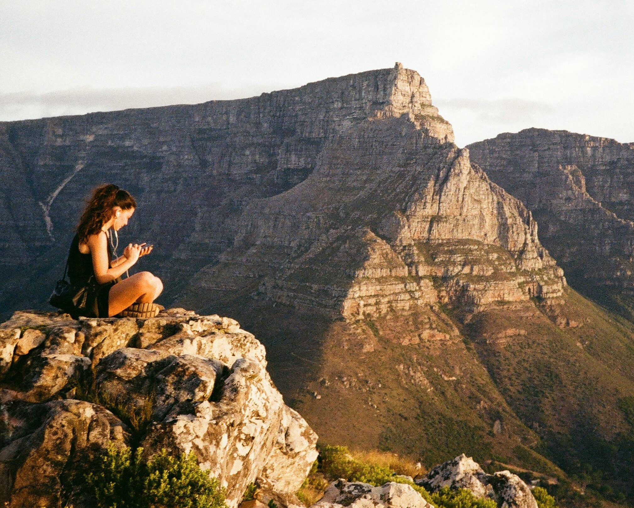 Woman looking at phone while seated atop Lion's Head with Table Mountain in background, Cape Town best photography spots Woman looking at phone while seated atop Lion's Head with Table Mountain in background, Cape Town best photography spots