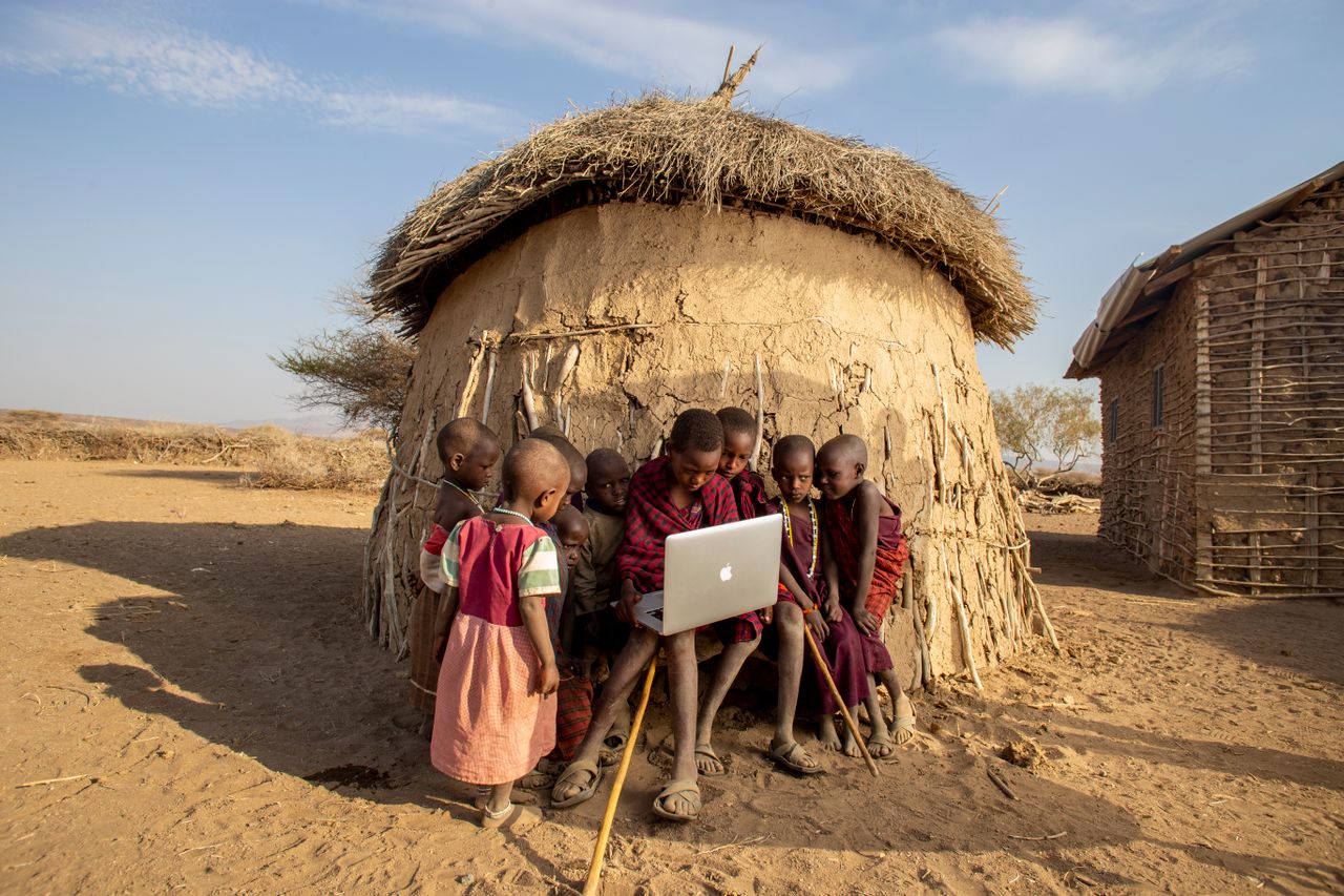 Masai children seated in front of mud hut looking at single Macbook