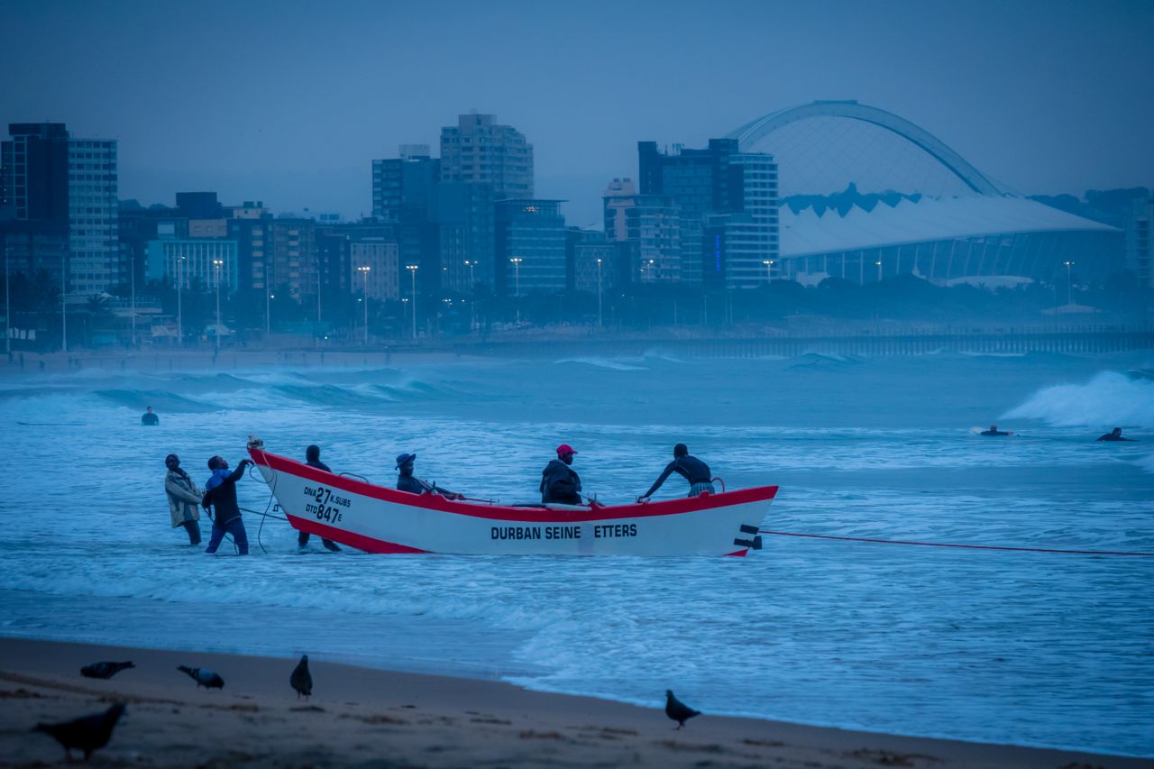 Fisherman in boat on Golden Mile of Durban with buildings and Moses Mabhida Stadium in background, dawn or dusk